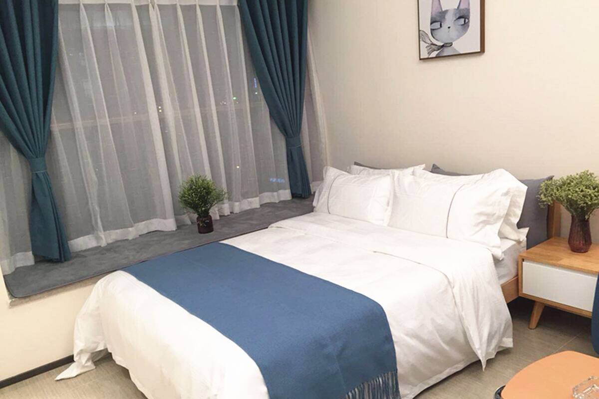 Una cama bien tendida con ropa de cama blanca y azul, una pequeña planta en maceta y un cuadro en la pared.
