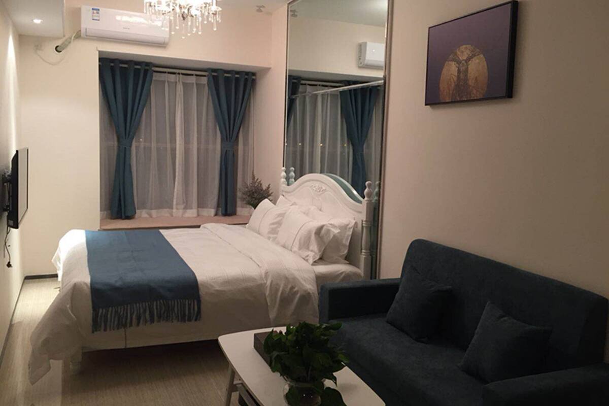 Un dormitorio con cama, una mesita de noche con una planta, un sofá de color oscuro, un espejo grande y un cuadro en la pared.