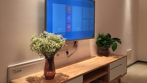 A modern living room with a flat-screen TV mounted on the wall, a wooden console table with a vase of flowers, and a potted plant. - Goodview Apartment Hotel (Changsha)