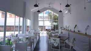 A spacious indoor dining area with high ceilings, large windows, and multiple ceiling fans. Tables are set with white tablecloths and chairs, and there are potted plants on a shelf along the wall. - Nan Nakara Boutique (Nan)