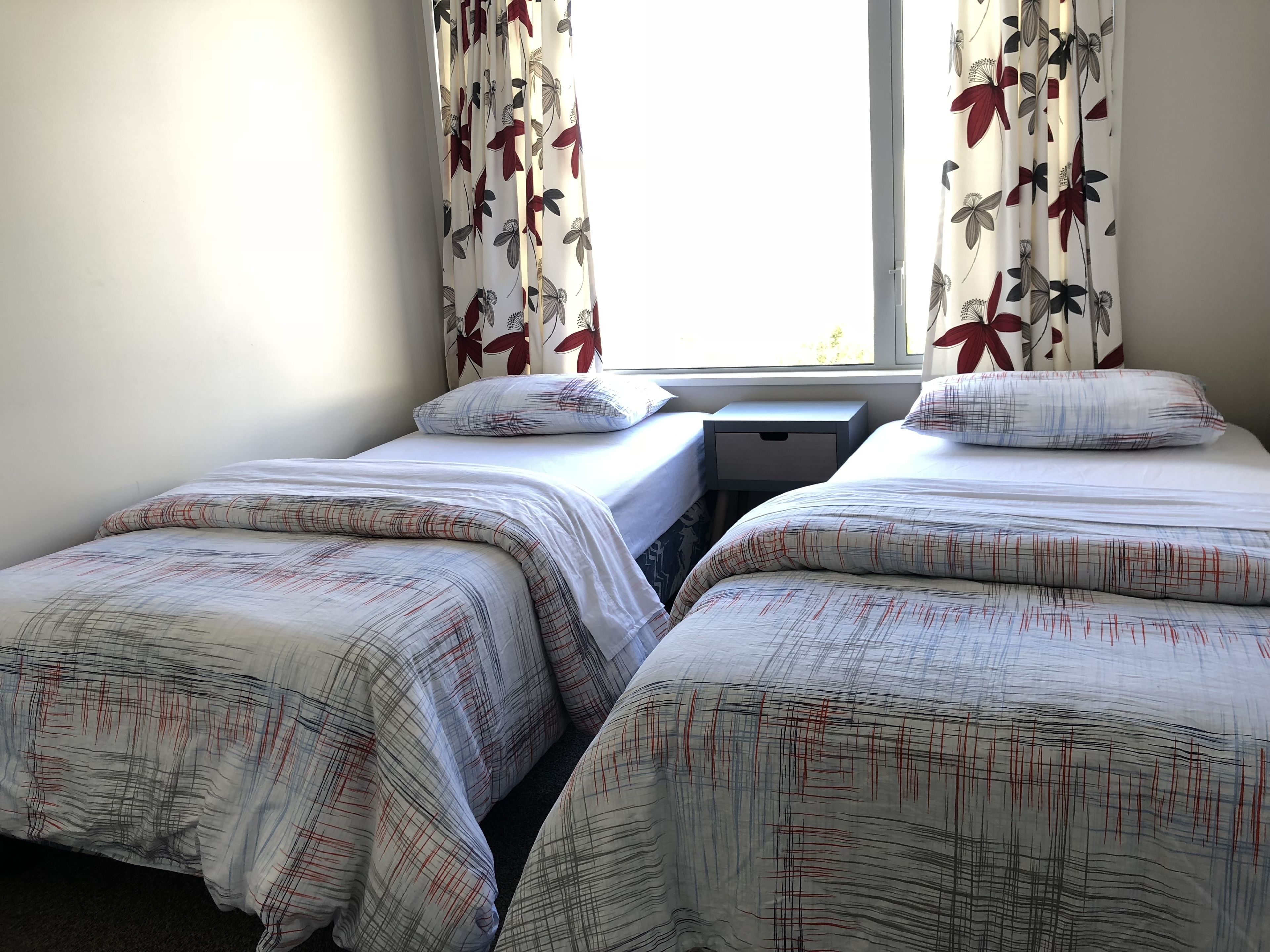 Ein Zimmer mit zwei Einzelbetten, einem Nachttisch und einem Fenster mit blumigem Vorhang.