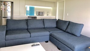 A modern living room with a grey sectional sofa, a white coffee table, and a kitchen area with a microwave and sink. - Lovely Sunny Home Sweet Home (Christchurch)