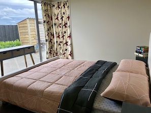Ein Schlafzimmer mit einem Bett, einem Fenster mit Vorhängen, einem kleinen Tisch und einem Stapel Bücher auf einem Nachttisch.
