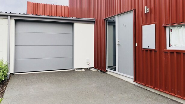 A red industrial building with a grey garage door, a glass door, and a small window with a curtain. - Lovely Sunny Home Sweet Home (Christchurch)