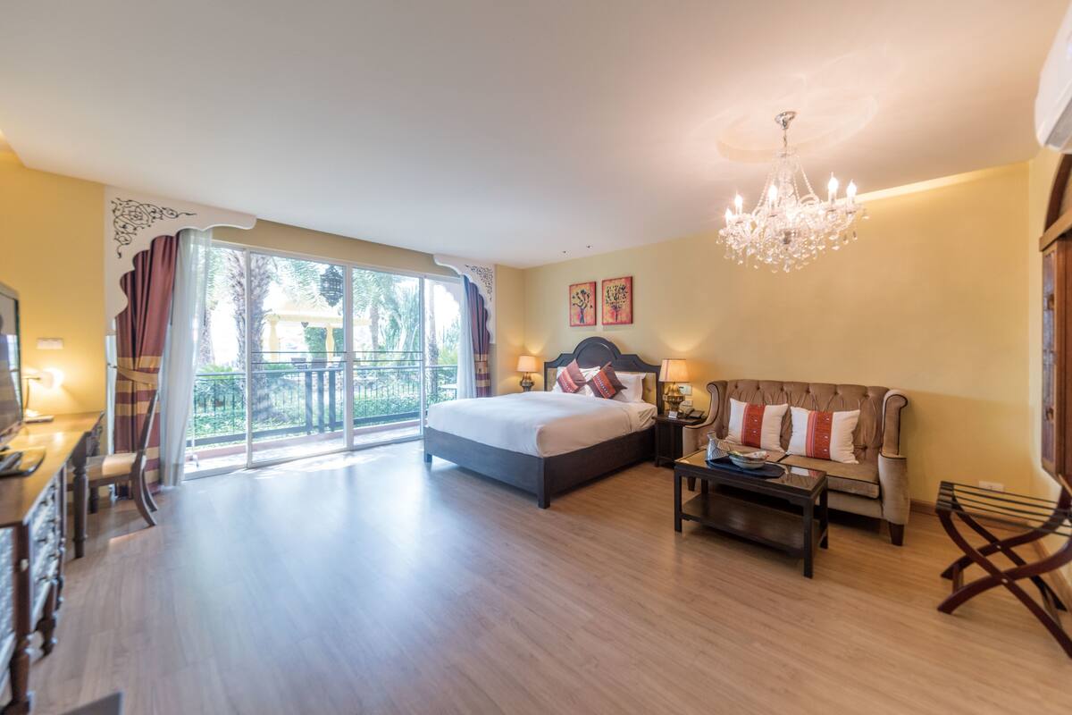 A spacious bedroom with a large bed, a sofa, a coffee table, and a chandelier. There is a balcony with curtains and a view of greenery.