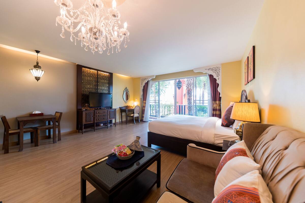 A hotel room with a bed, a sofa, a desk with a chair, a television, and a balcony with curtains.
