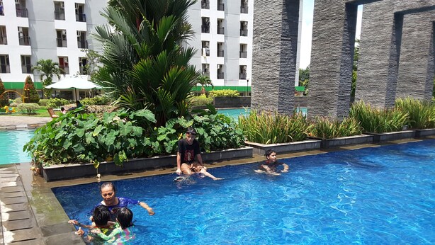 A swimming pool with clear blue water, surrounded by greenery and a building with balconies. - Green Lake View Luxury Apartment by Indah (South Tangerang)