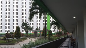 A corridor with a view of a high-rise building, a covered walkway, and a small garden area with plants and a bench. - Green Lake View Luxury Apartment by Indah (South Tangerang)