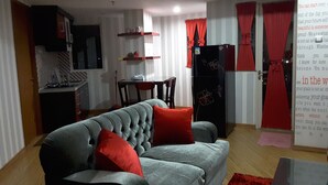 A compact living space with a grey sofa, red cushions, a dining table, and a kitchen area with a refrigerator and shelves. - Green Lake View Luxury Apartment by Indah (South Tangerang)