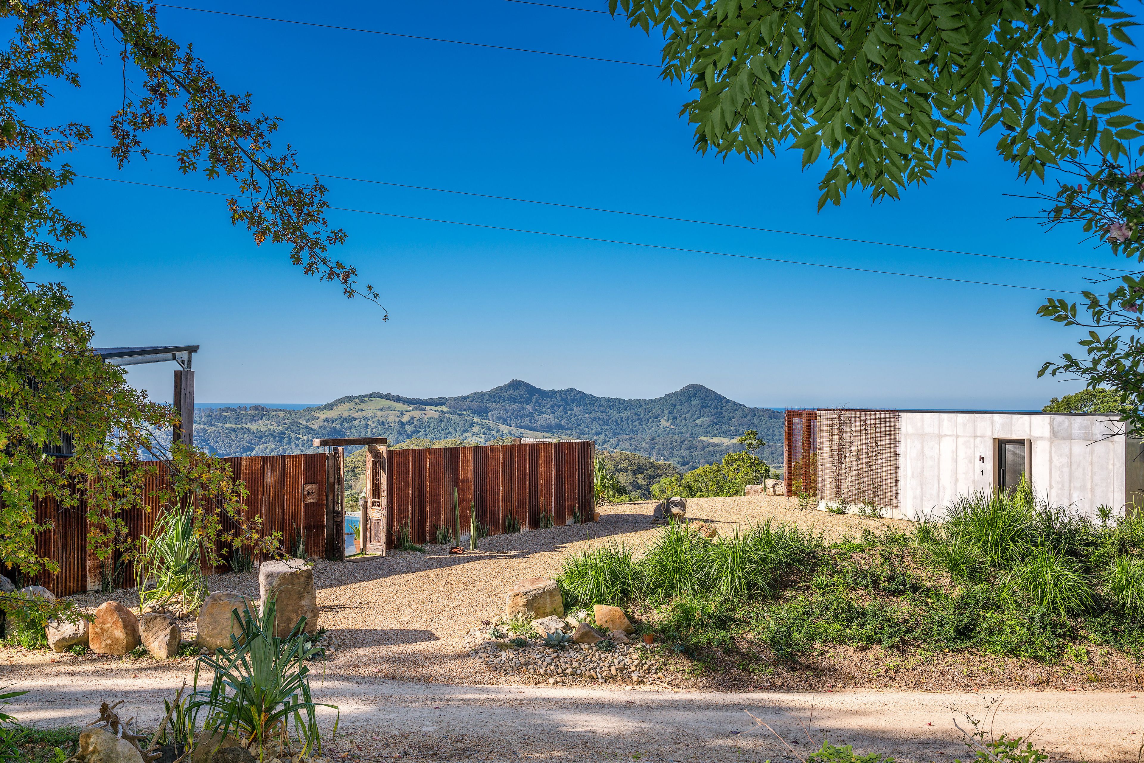 A rustic cabin with a gravel driveway, surrounded by natural vegetation and a mountainous backdrop.