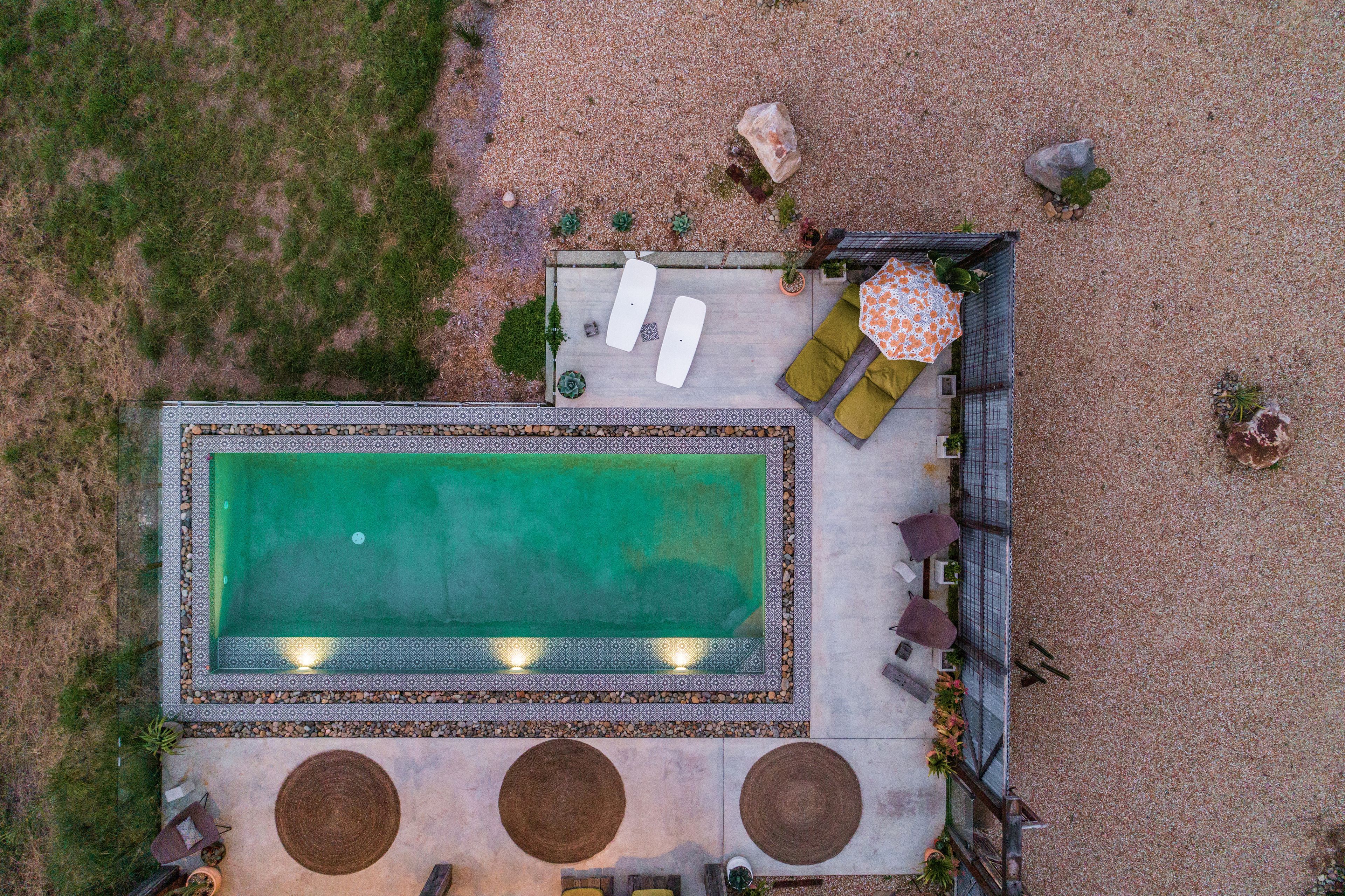 Vista aérea de un área de piscina en la azotea, con una piscina rectangular, dos sillones de descanso, una mesa pequeña con un paraguas estampado y plantas alrededor.