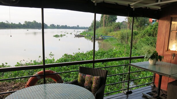 Una habitación con un ventanal grande con vista a un río tranquilo, muebles de mimbre y una pequeña mesa con una planta en maceta.