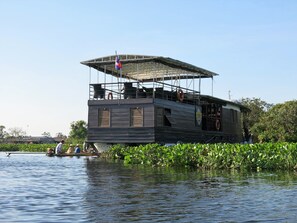 Une péniche dotée d’un pont supérieur couvert, entourée d’eau et de verdure.