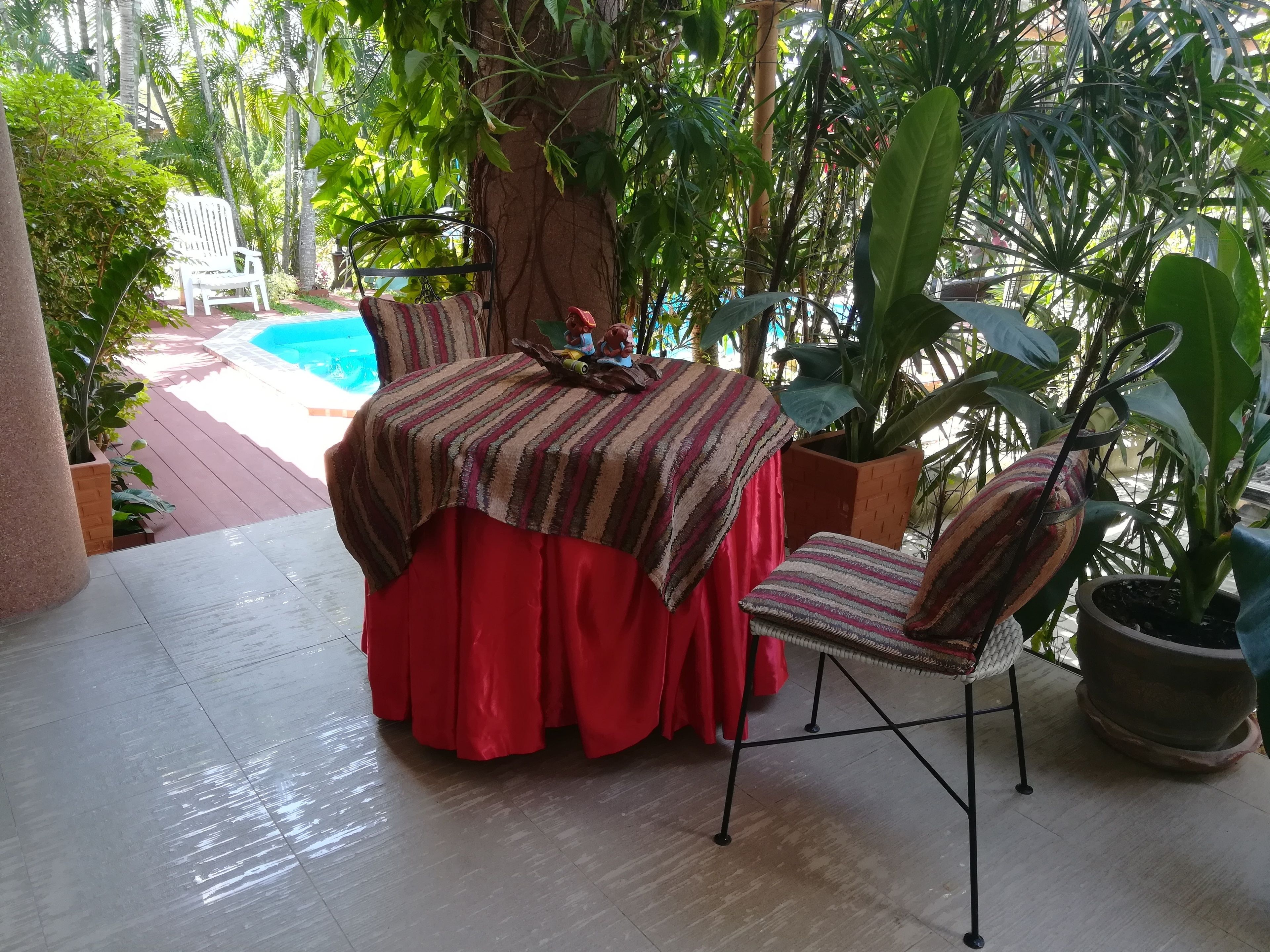 A covered outdoor seating area with a table and chairs, surrounded by lush greenery and a view of a swimming pool.