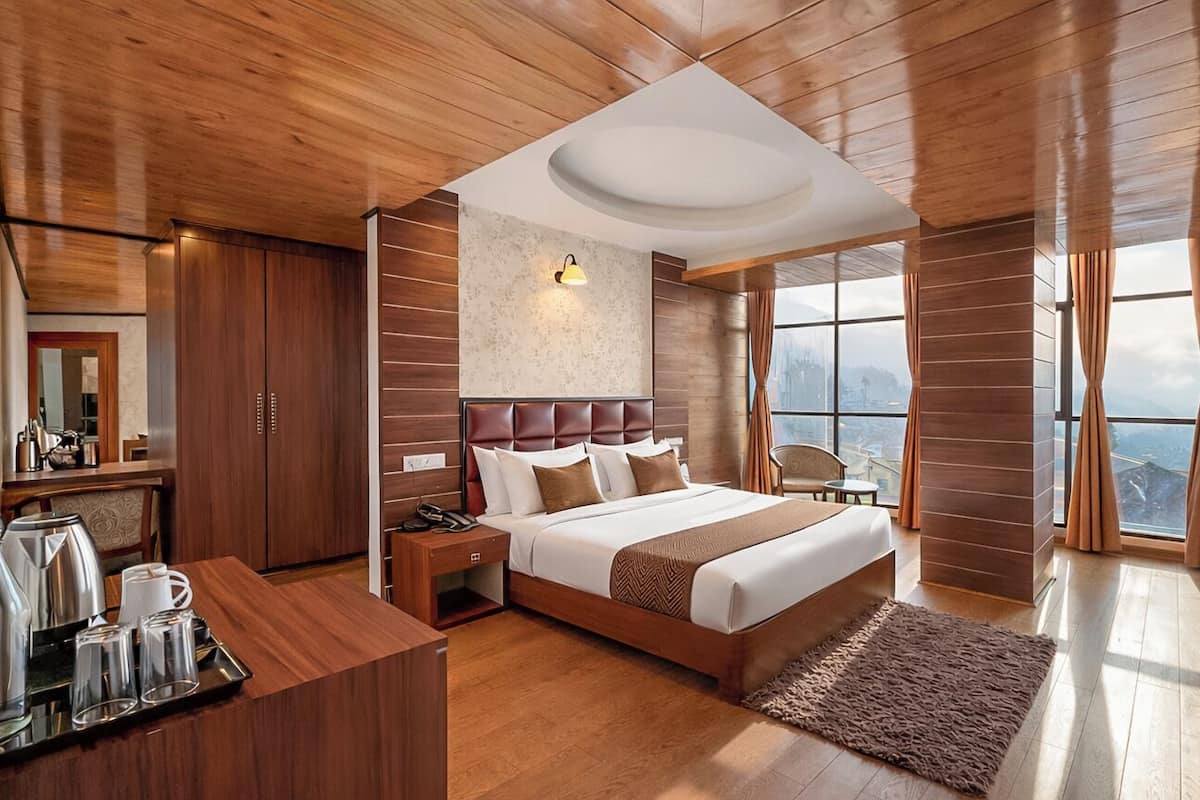 Habitación de hotel moderna con techo de madera, cama grande con cabecera, escritorio con equipo para preparar café y vistas de la ciudad a través de amplias ventanas.