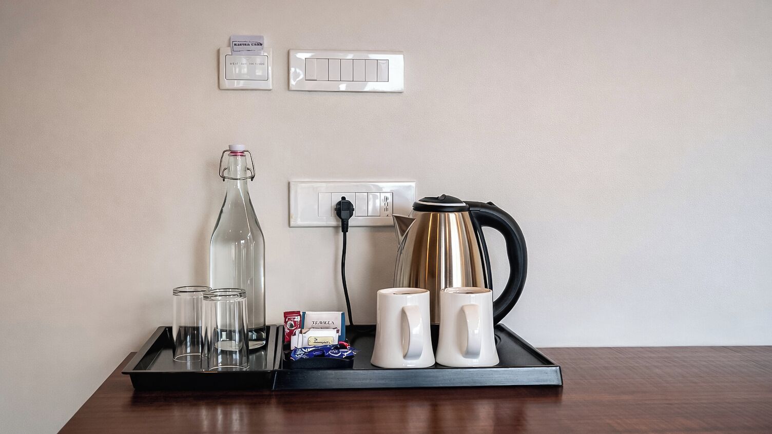 A tray with a glass bottle, two glasses, a mug, a kettle, and a box of tissues on a wooden table.