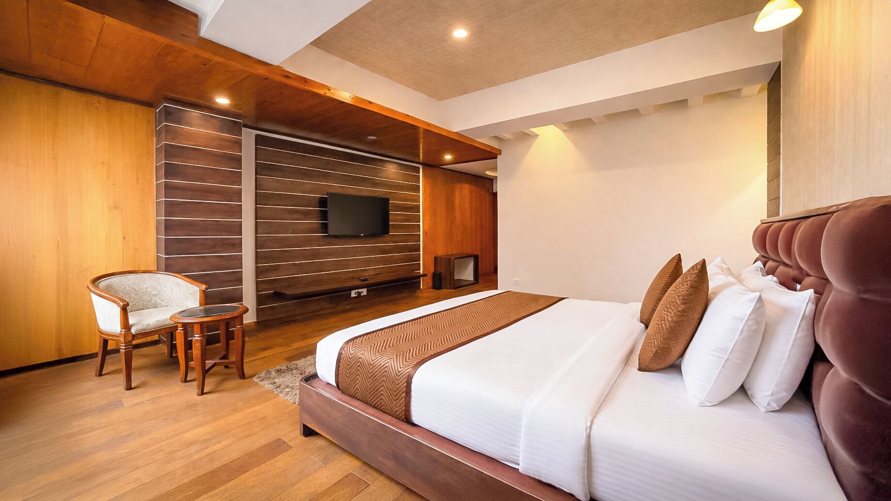 Une chambre moderne avec un grand lit, un téléviseur fixé au mur et un coin salon comprenant un fauteuil et une petite table.
