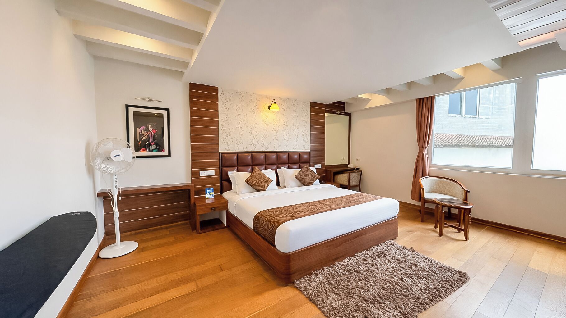 A modern hotel room with a large bed, a bedside table, a chair, a standing fan, and a window with curtains.