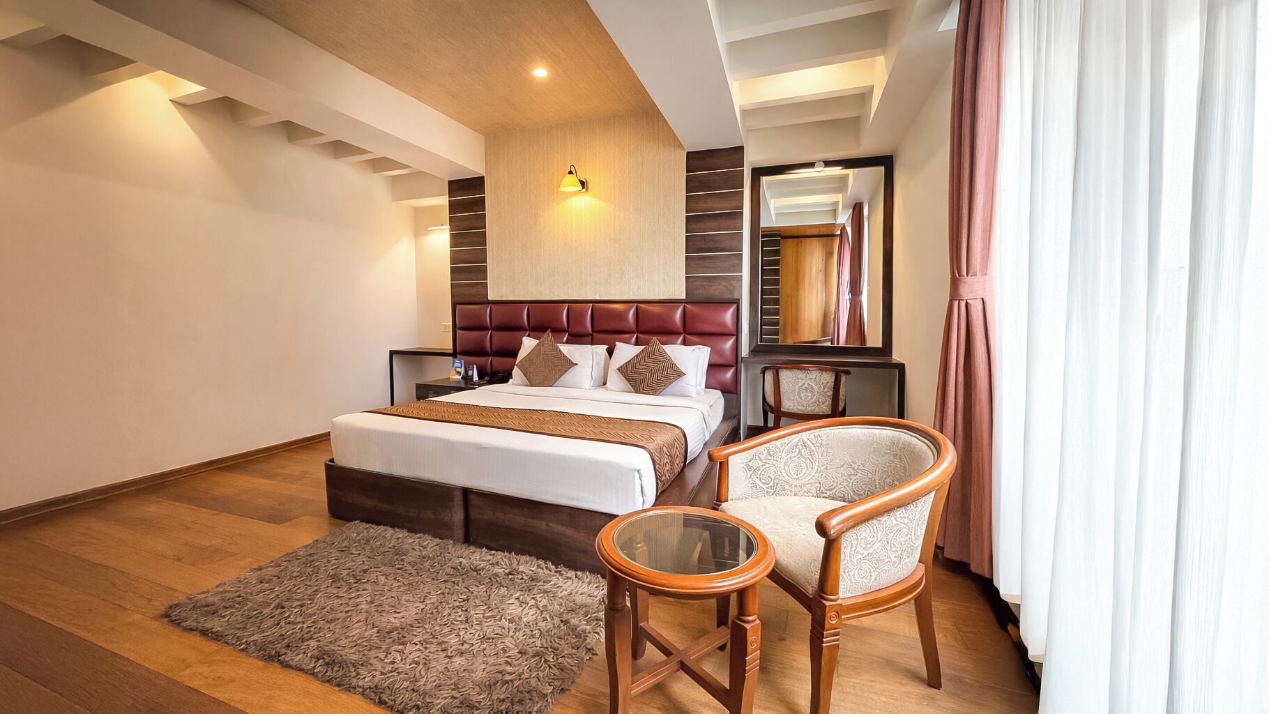 Quarto de hotel com cama, cadeira, mesa pequena, espelho e janela com cortinas.
