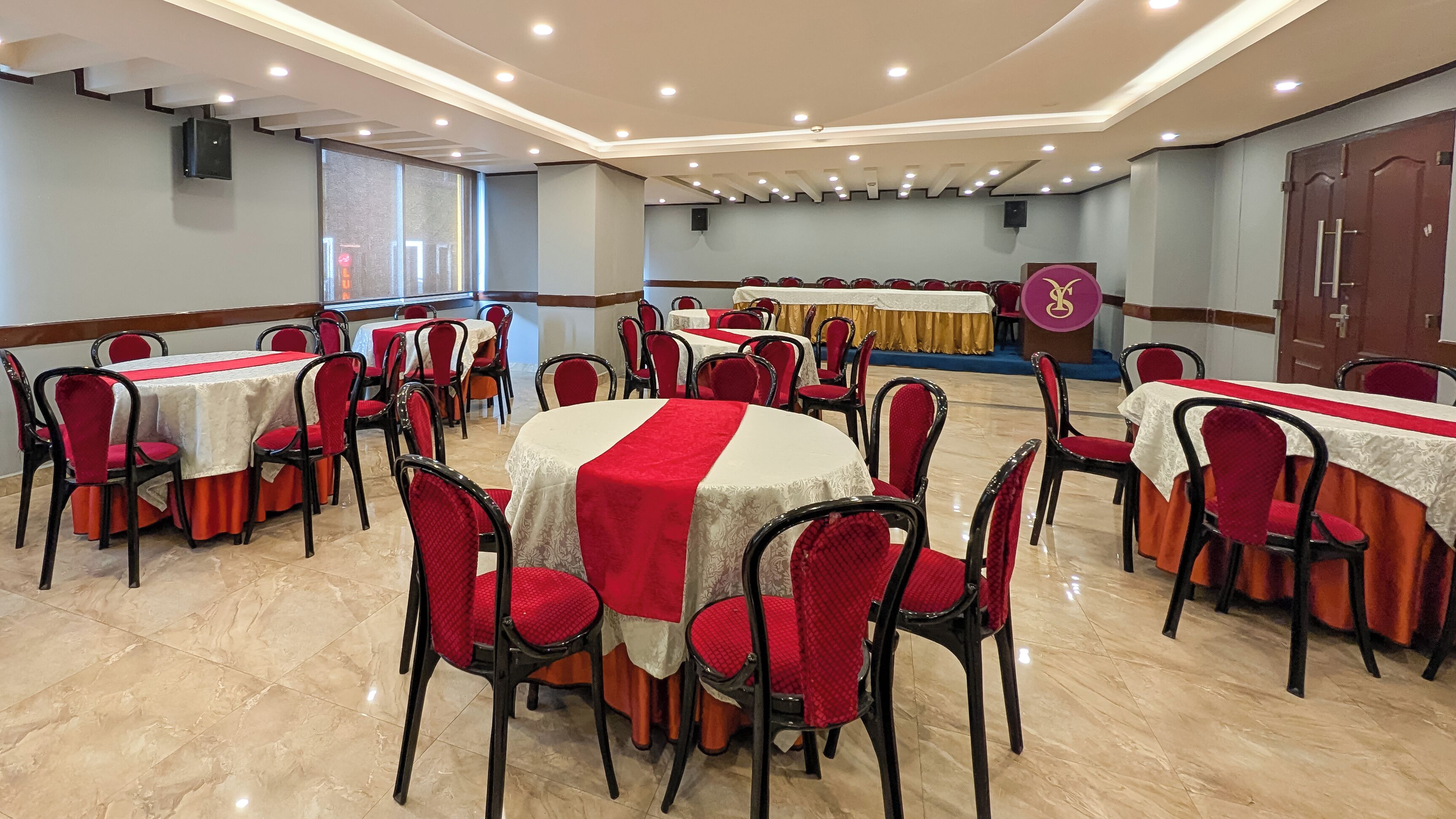 Un salón de conferencias con mesas redondas, sillas rojas y un escenario con un atril.