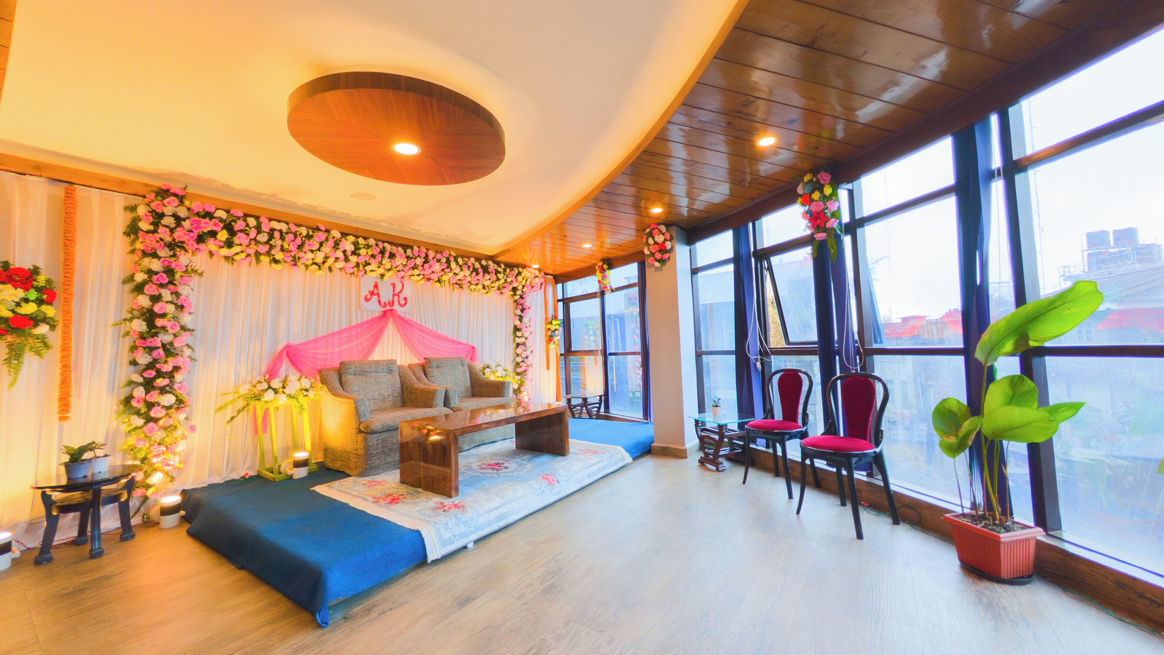 A spacious room with a wooden ceiling, large windows, a sofa, chairs, and potted plants.