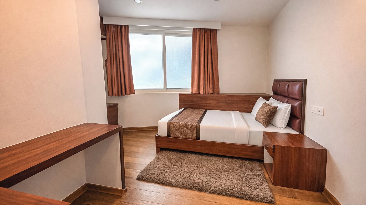 A bedroom with a wooden bed, a nightstand, a desk, and a window with curtains.