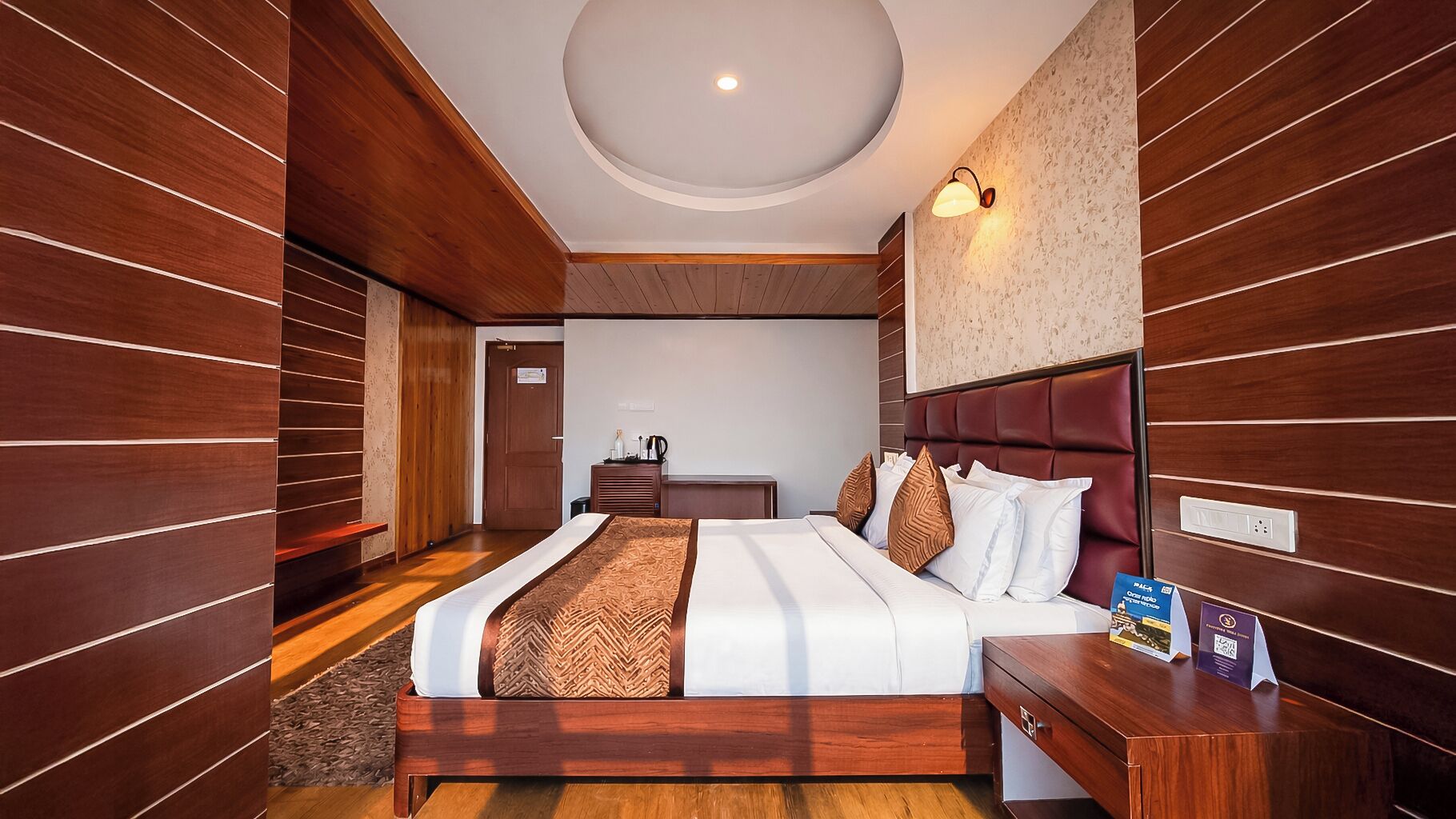 Une chambre d’hôtel avec un décor de panneaux en bois, un grand lit avec de nombreux oreillers, une table de chevet et un bureau avec une lampe.