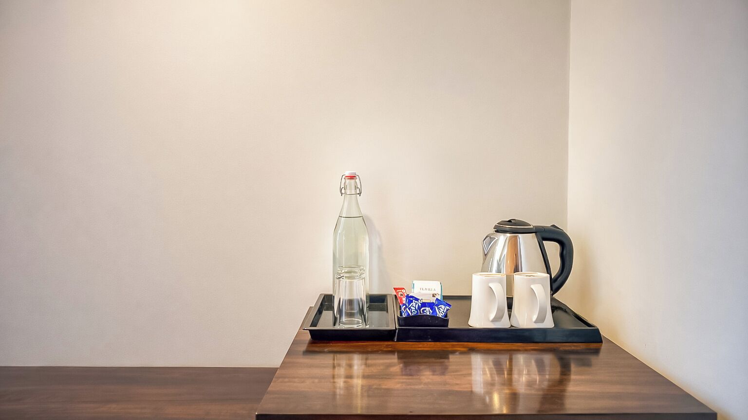 Habitación de hotel con una mesa de madera, una botella de vidrio, dos tazas blancas, una tetera y una bandeja con sobres.