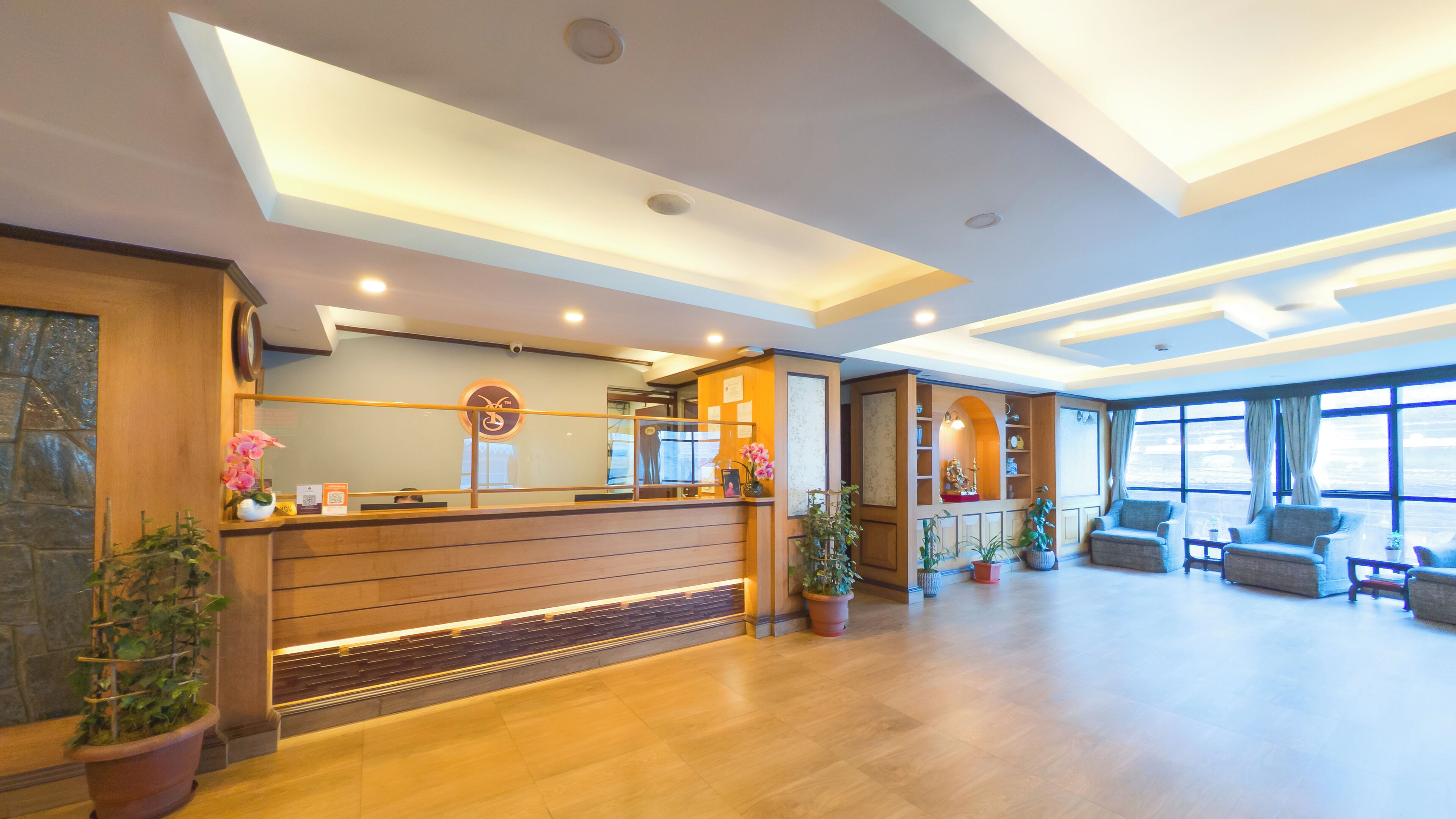 A hotel reception area with a wooden reception desk, potted plants, and a seating area with sofas and a coffee table.