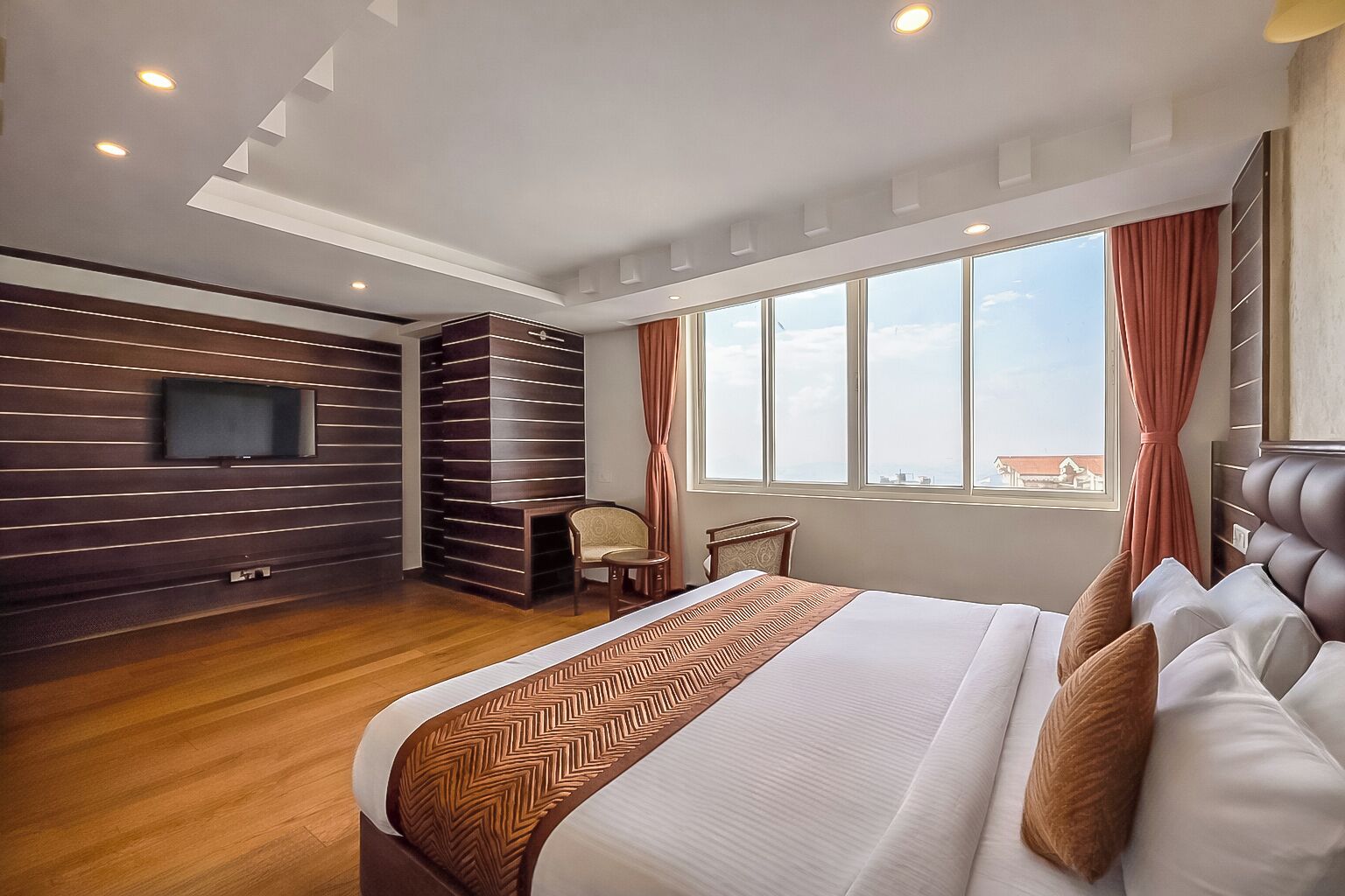 A modern hotel room with a large bed, a TV mounted on the wall, a seating area with a chair and table, and a large window with curtains.