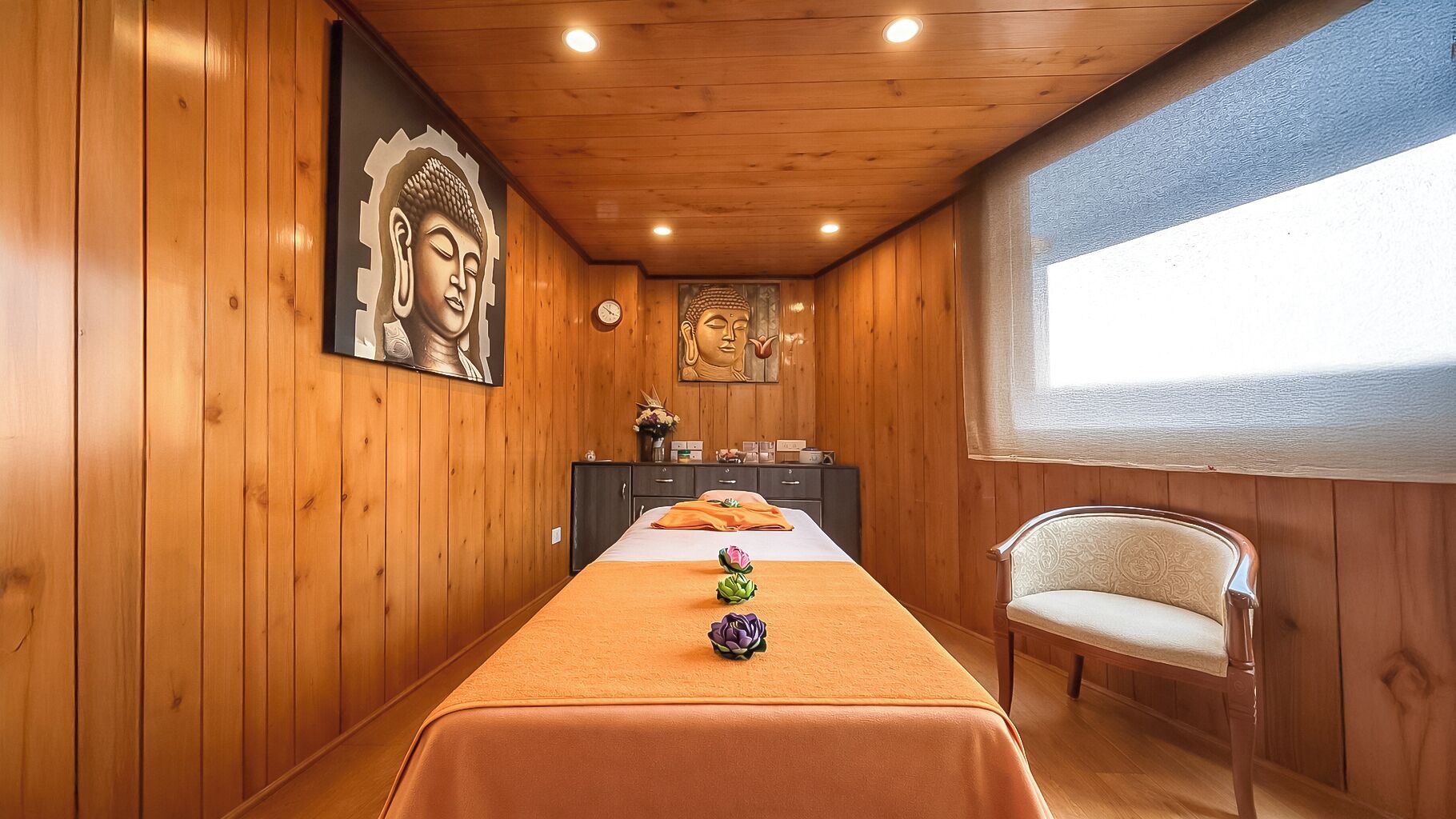 A room with wooden paneling, a bed with an orange bedspread, a chair, and two Buddha statues on the wall.