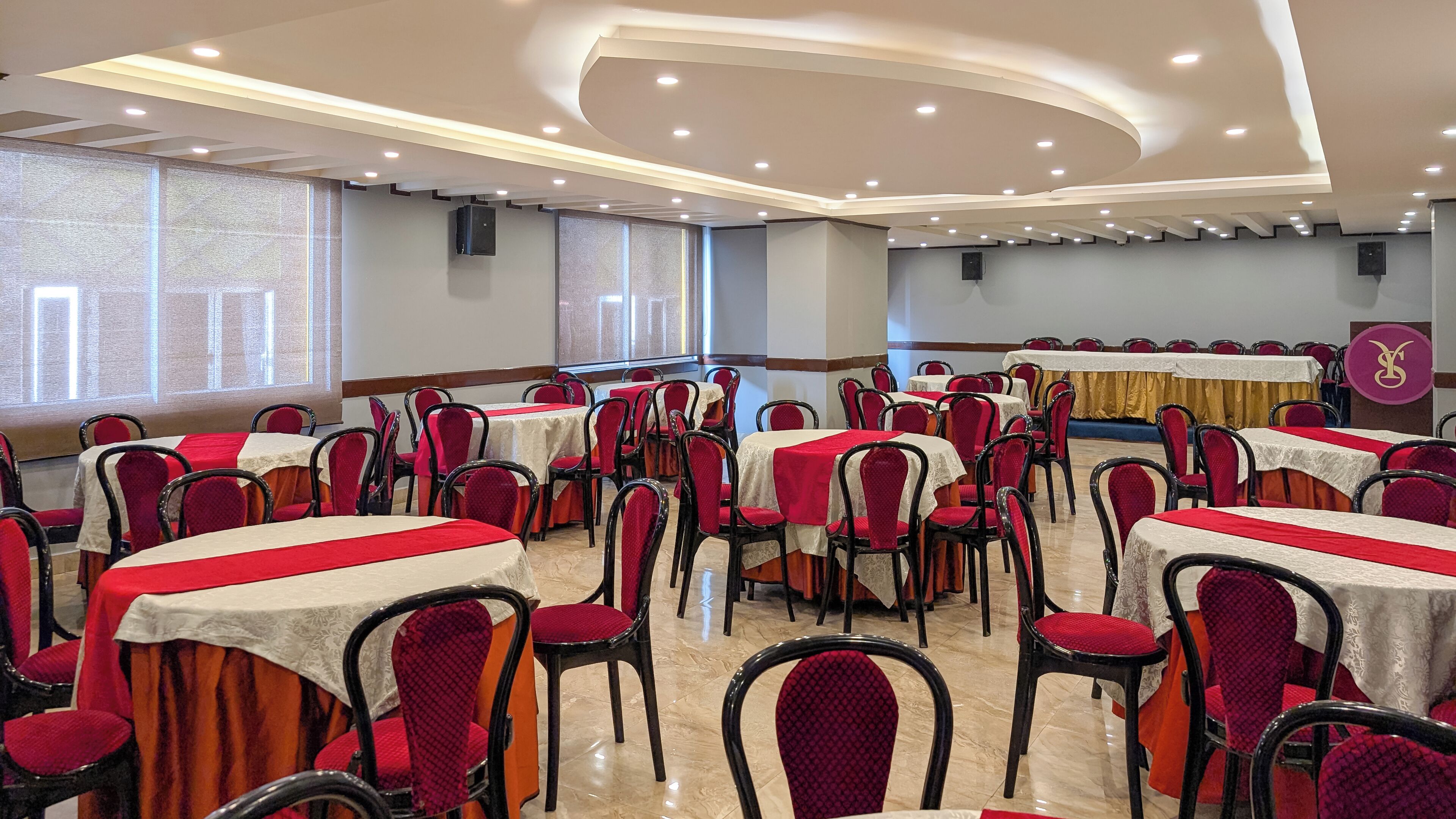 Un salón de conferencias con mesas y sillas redondas, equipado con luces de techo y una pantalla para proyector.