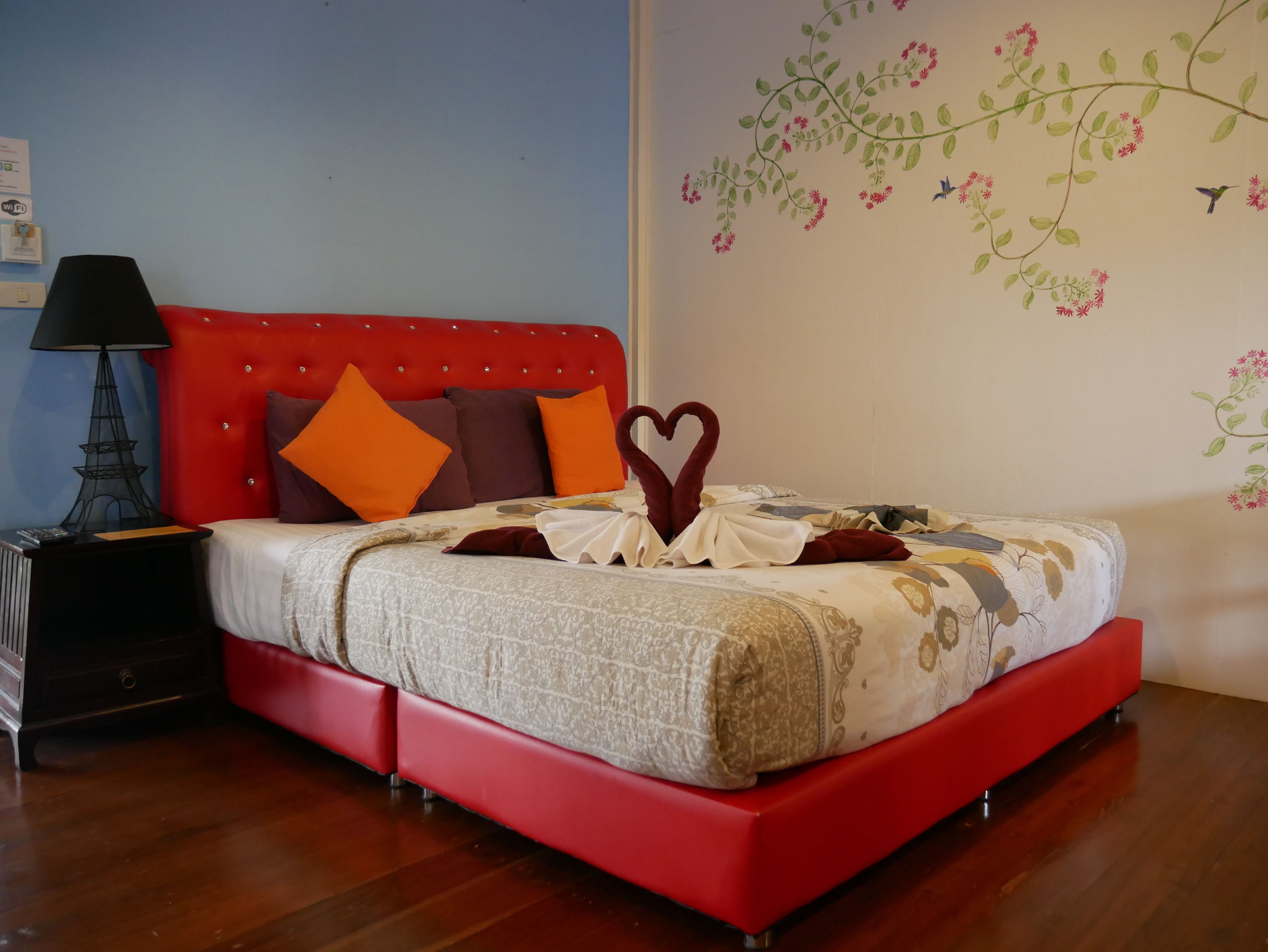 Una cama bien hecha con un cabecero rojo, una decoración de toalla con forma de corazón y un mural floral en la pared.