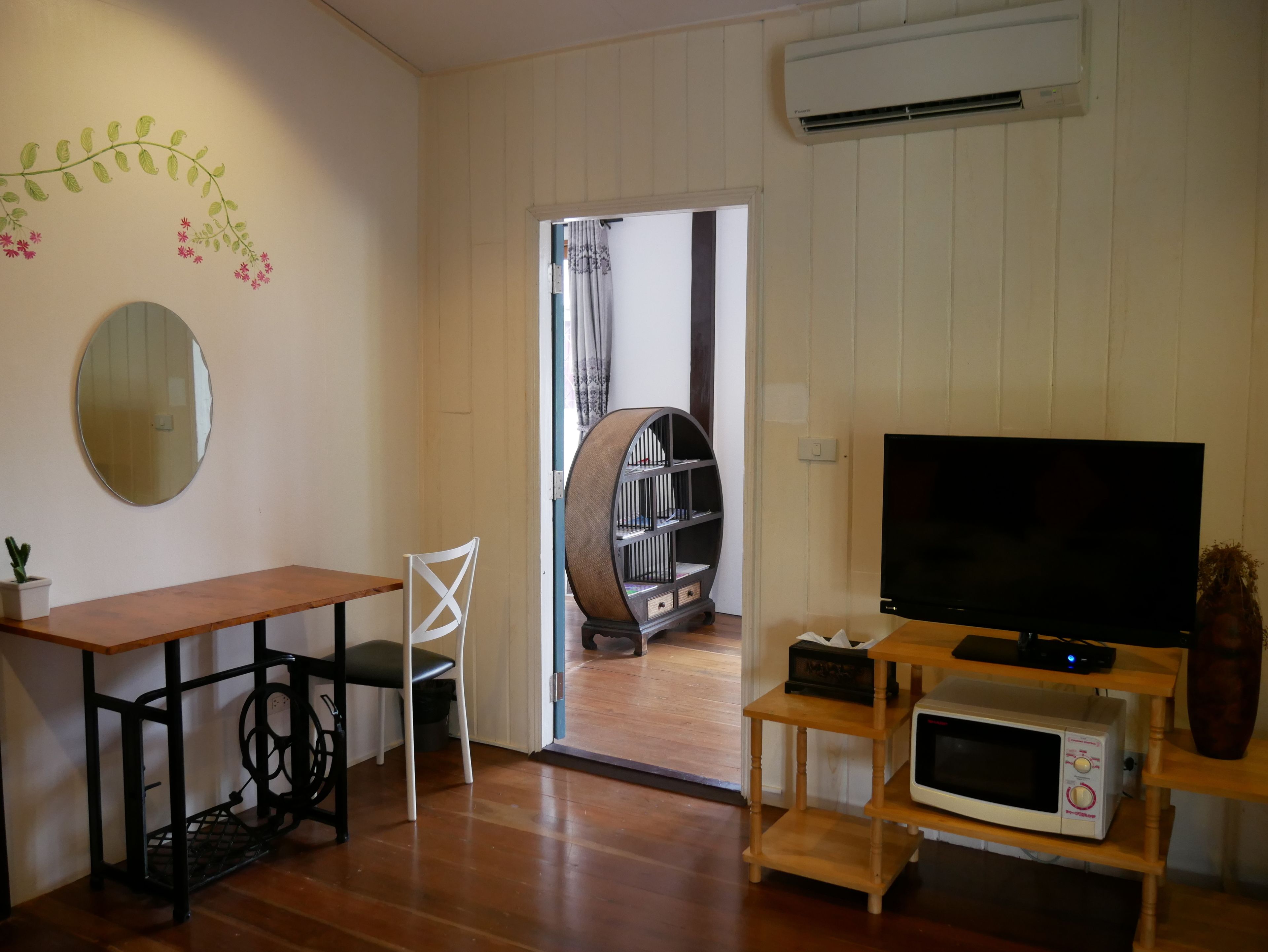 Una sala de estar con una mesa de madera, una silla blanca, un televisor, una estantería y un equipo de aire acondicionado.