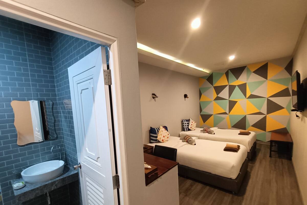 Habitación de hotel con baño que presenta un muro de azulejos azules, un lavamanos blanco y un espejo. La habitación tiene dos camas con ropa blanca y almohadas decorativas, una mesita de noche de madera y un muro con diseño geométrico.