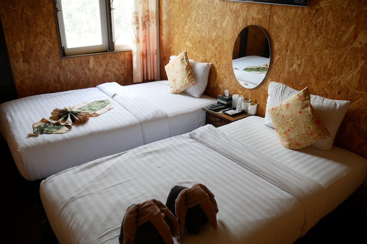 Habitación de hotel con dos camas, cabecero de madera, mesita de noche con espejo y ventana con cortinas.