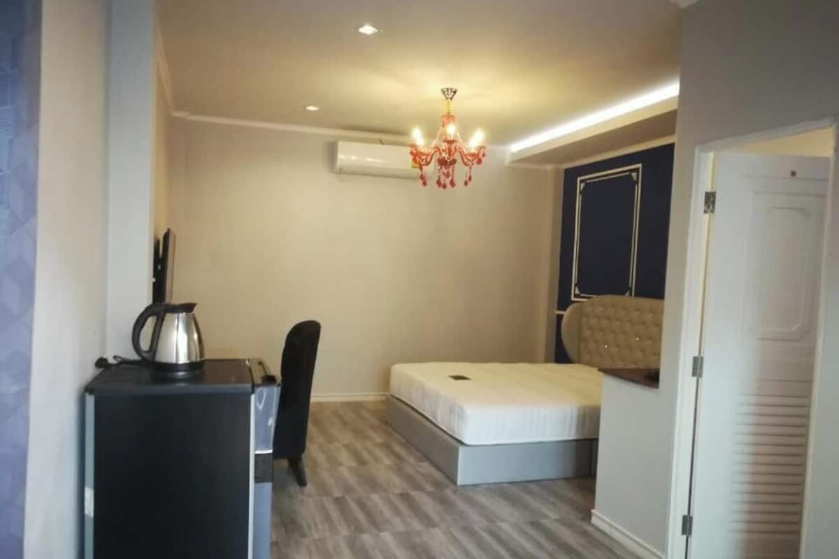 Una habitación pequeña y bien iluminada con una cama, un escritorio con un hervidor, una silla y una puerta.