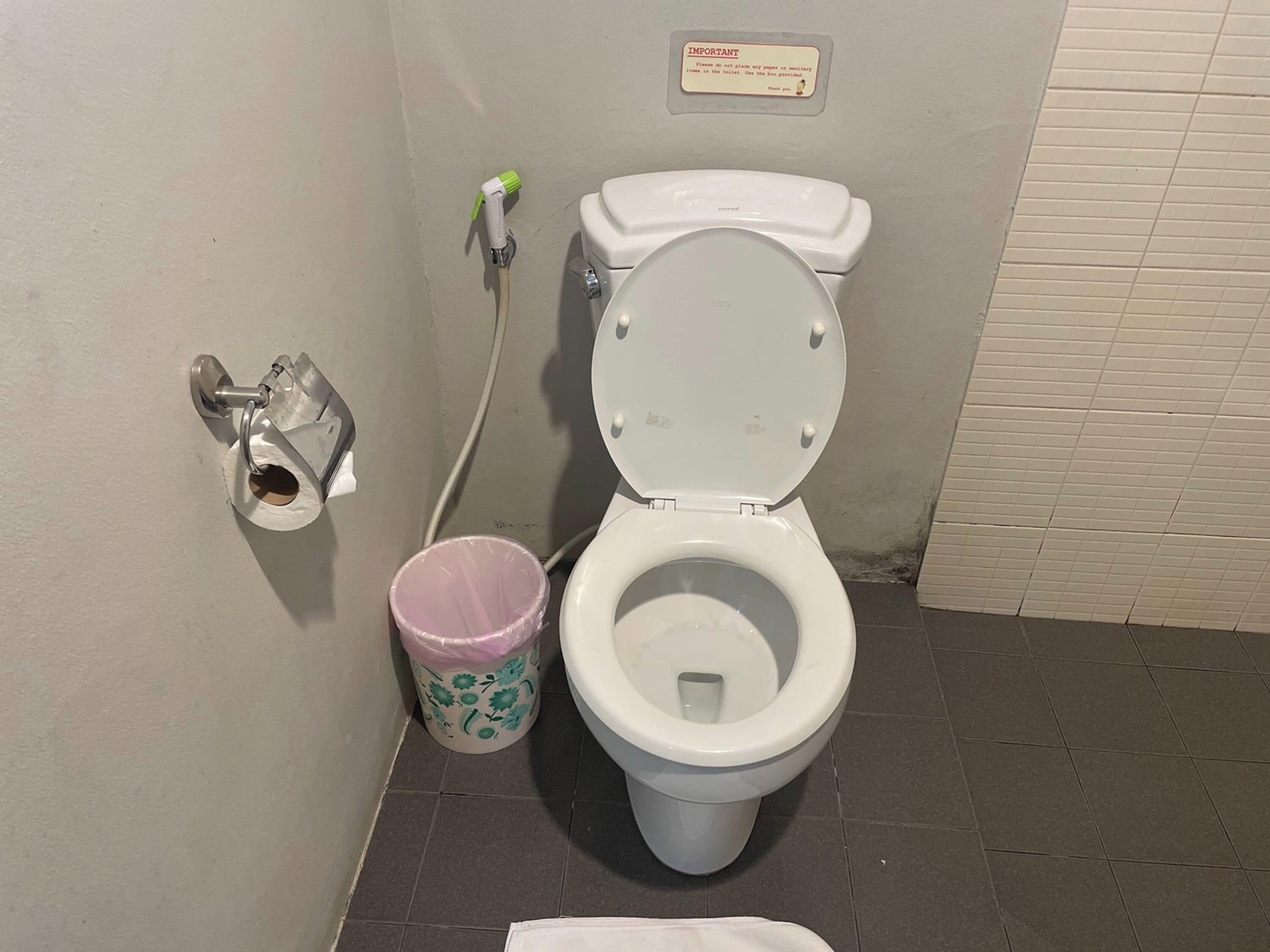 Un inodoro con asiento y tapa blancos, un cubo de basura rosa con motivos florales, un dispensador de papel montado en la pared y un letrero con un mensaje.