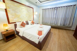 A neatly made bed with white and orange bedding, a wooden nightstand with a lamp, a stone-patterned wall, and a window with curtains. - Romantic Angkor Residence (Siem Reap)