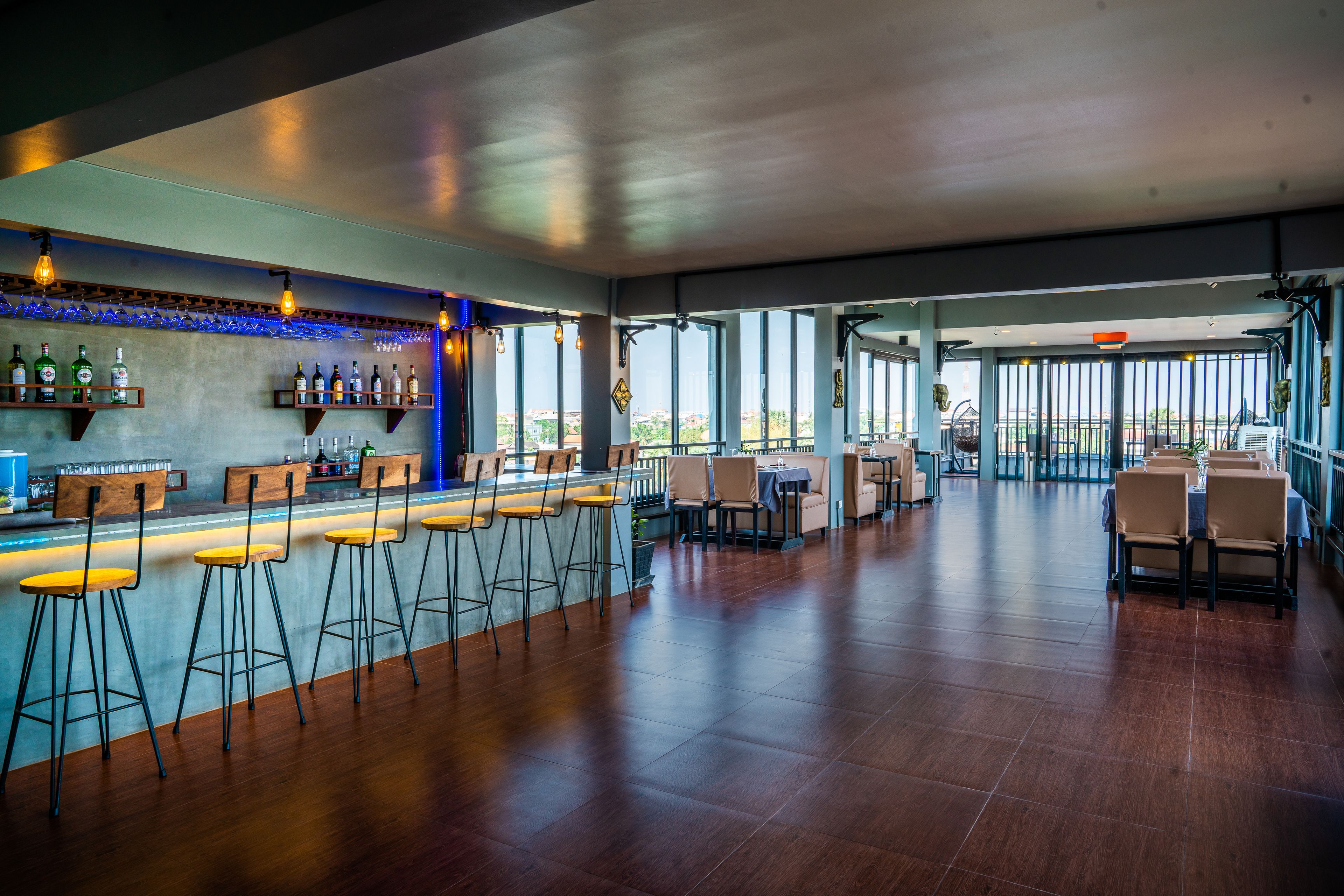 Un espace bar avec des tabourets et des étagères remplies de bouteilles, un espace repas avec des tables et des chaises, et de grandes fenêtres offrant une vue panoramique.