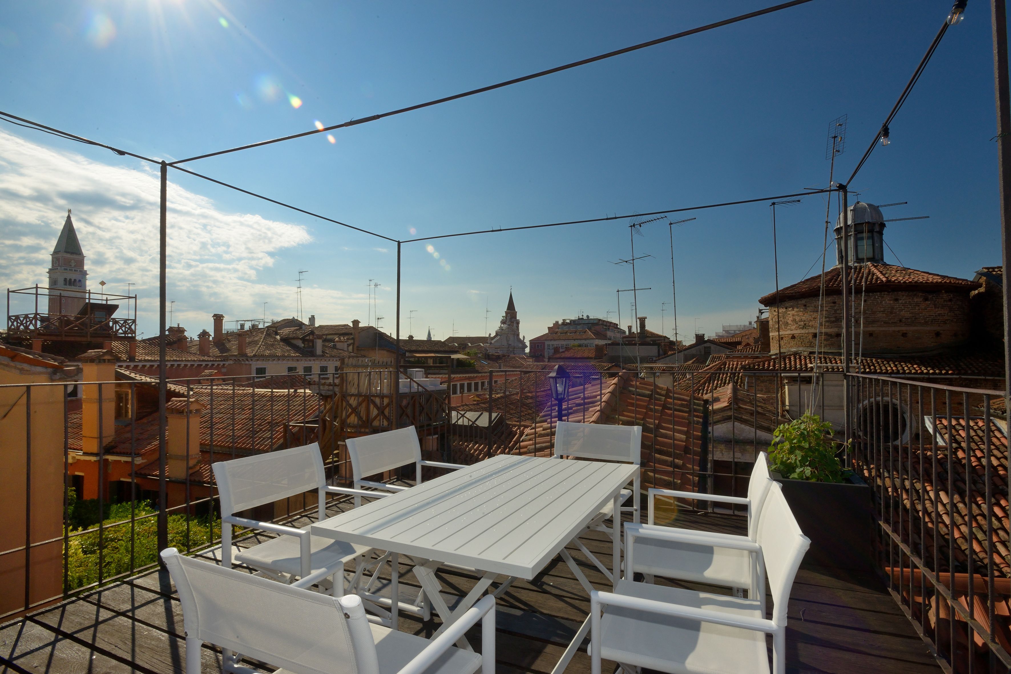 Een terras buiten met een witte tafel en stoelen, omringd door een metalen hek, met uitzicht op een stadsgezicht met historische gebouwen en een heldere lucht.