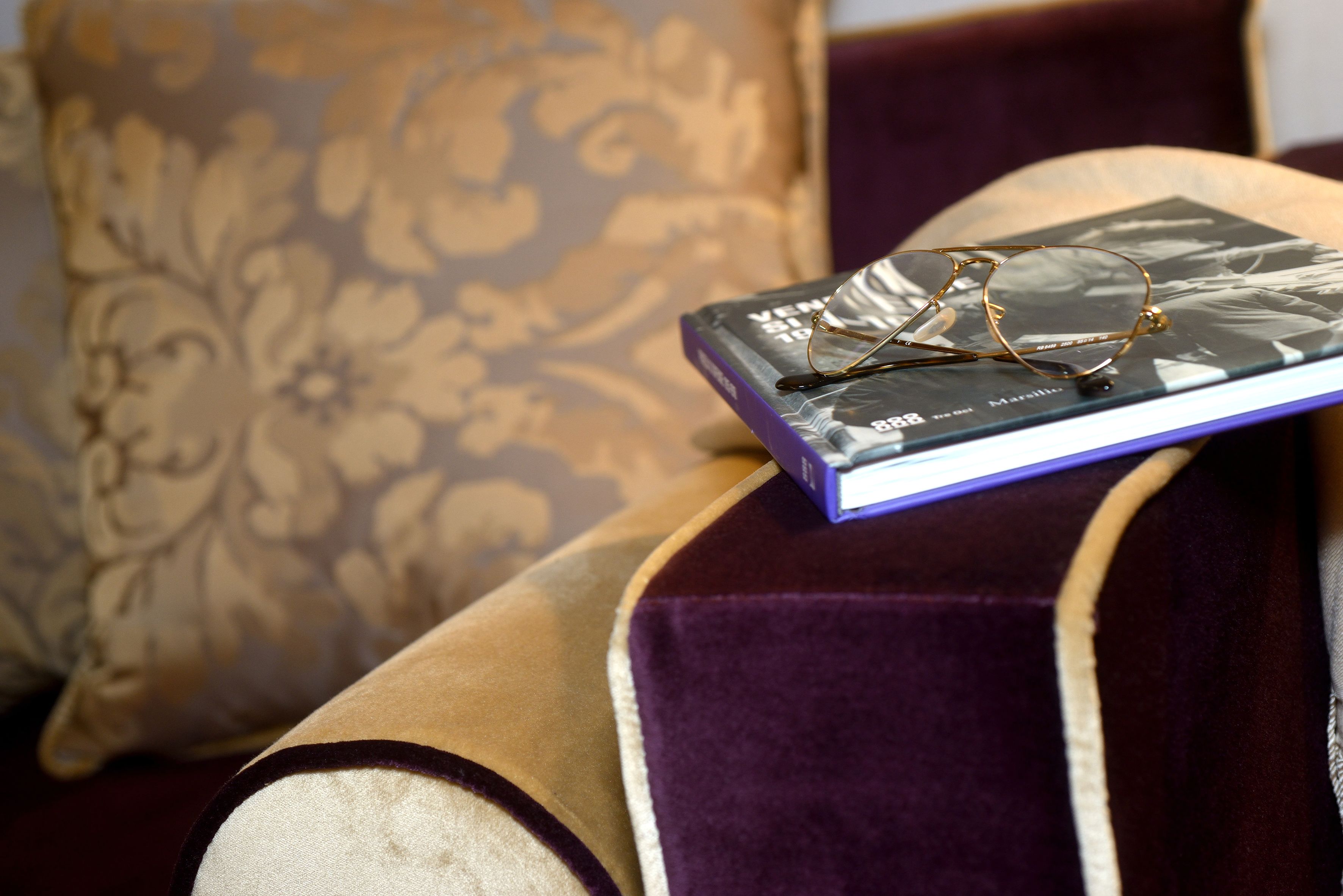 A pair of glasses on a book with a purple cover, placed on a cushioned surface with a patterned pillow in the background.