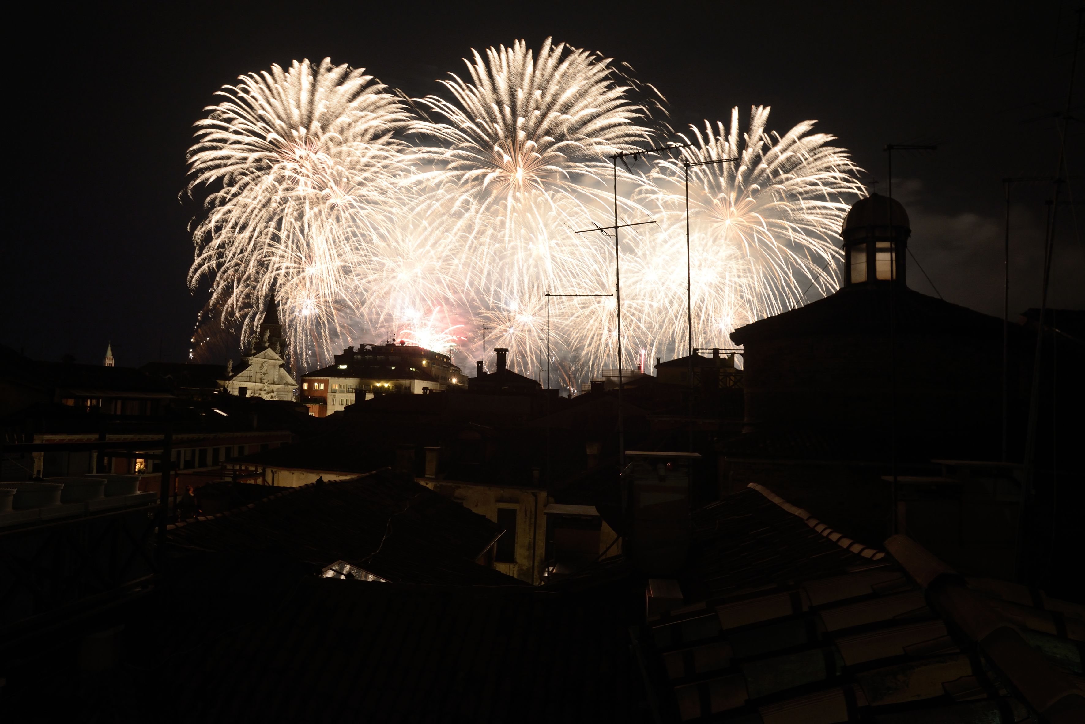 A nighttime view of a cityscape with buildings and a large display of fireworks in the sky.