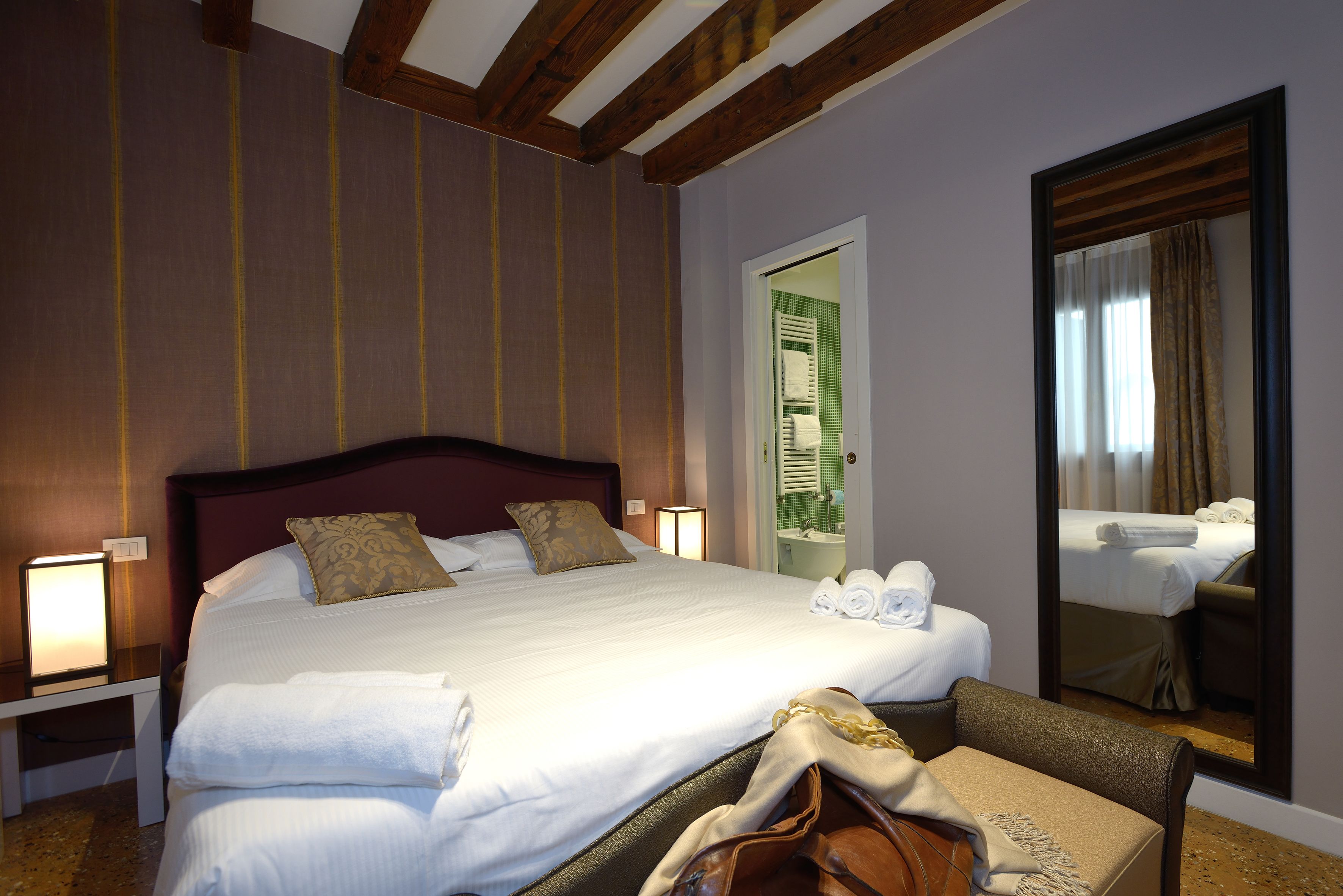 Ein Hotelzimmer mit einem großen Bett, Nachttischlampen, einem Spiegel und Blick ins Badezimmer durch eine Tür.