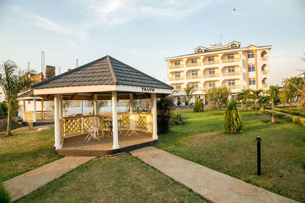 A gazebo with a tiled roof and the name "TSAVO" on its side, surrounded by a well-maintained lawn and a multi-story building in the background.