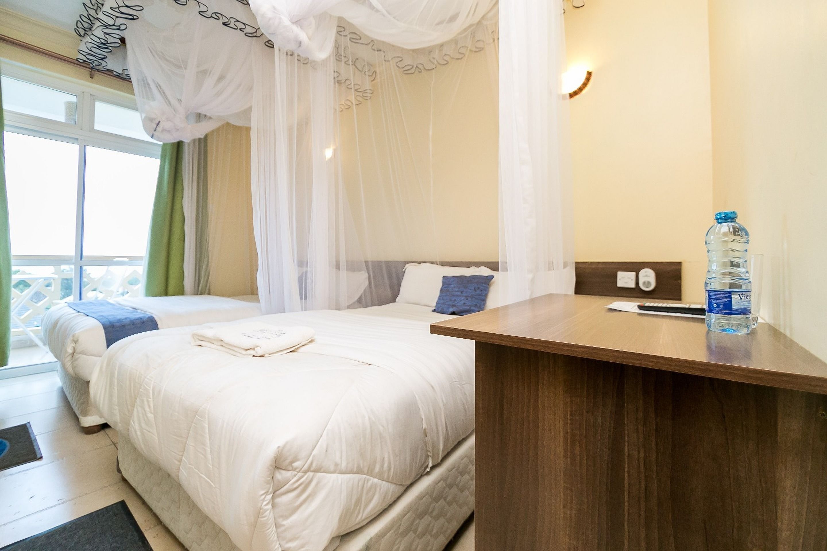 A hotel room with two beds, a wooden desk, a water bottle, and a window with curtains.