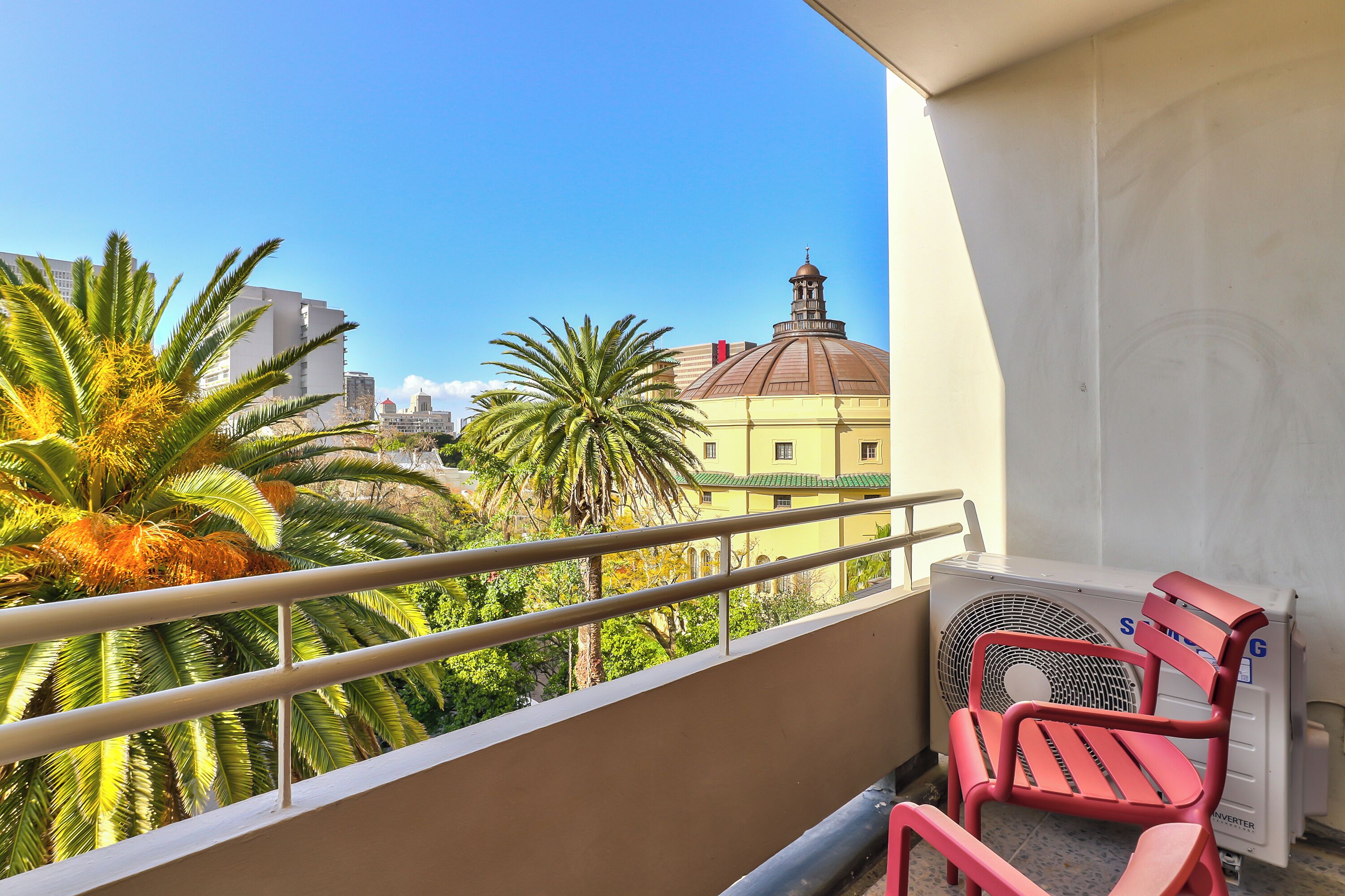 Un balcón con una silla roja, baranda blanca y vistas a palmeras y edificios.