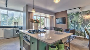 A modern kitchen with a central island, built-in appliances, and a dining area with a tropical-themed wall. - 437 St Martini Gardens (Cape Town)