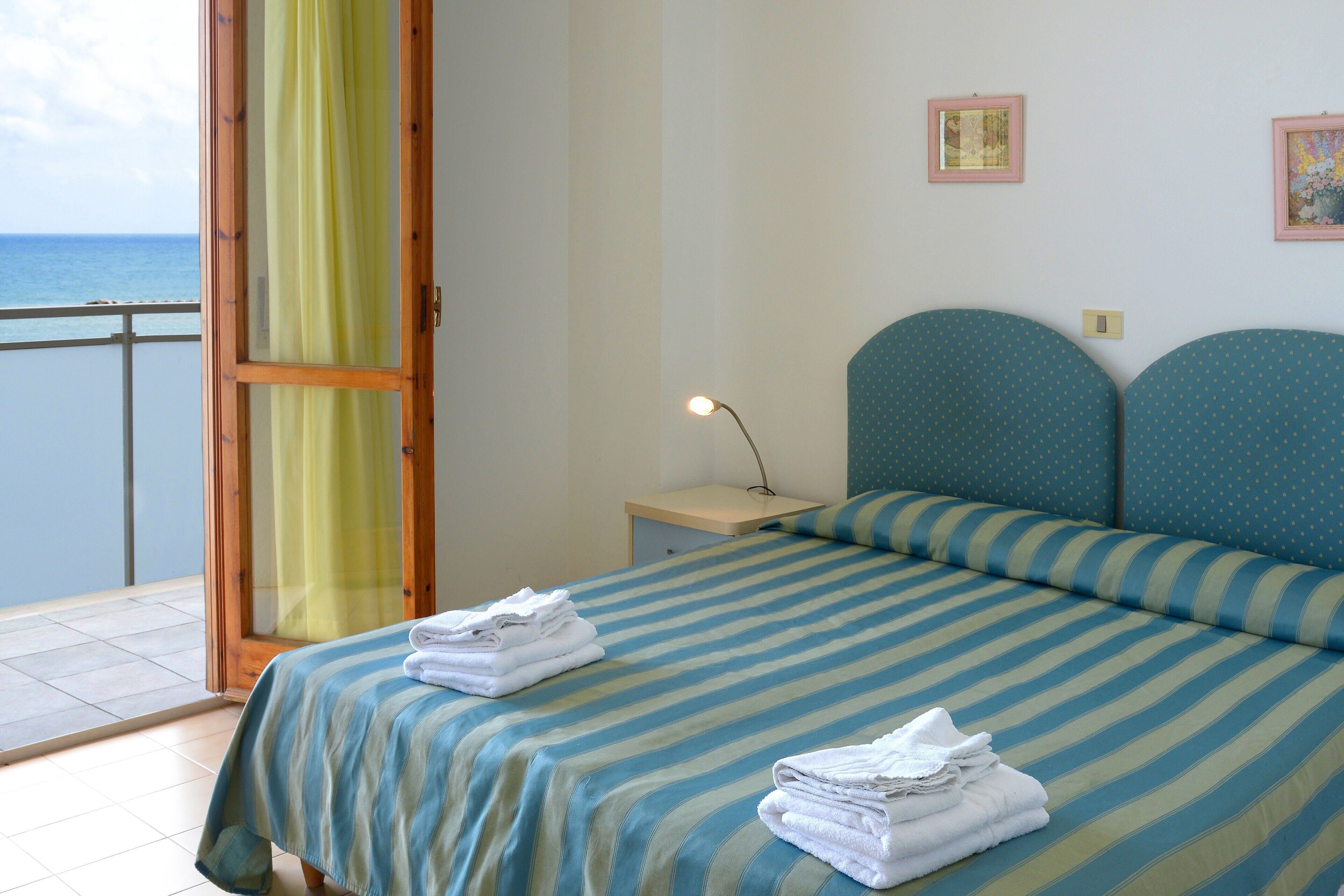 A hotel room with a bed, a nightstand, a lamp, and a view of the sea through a curtain.