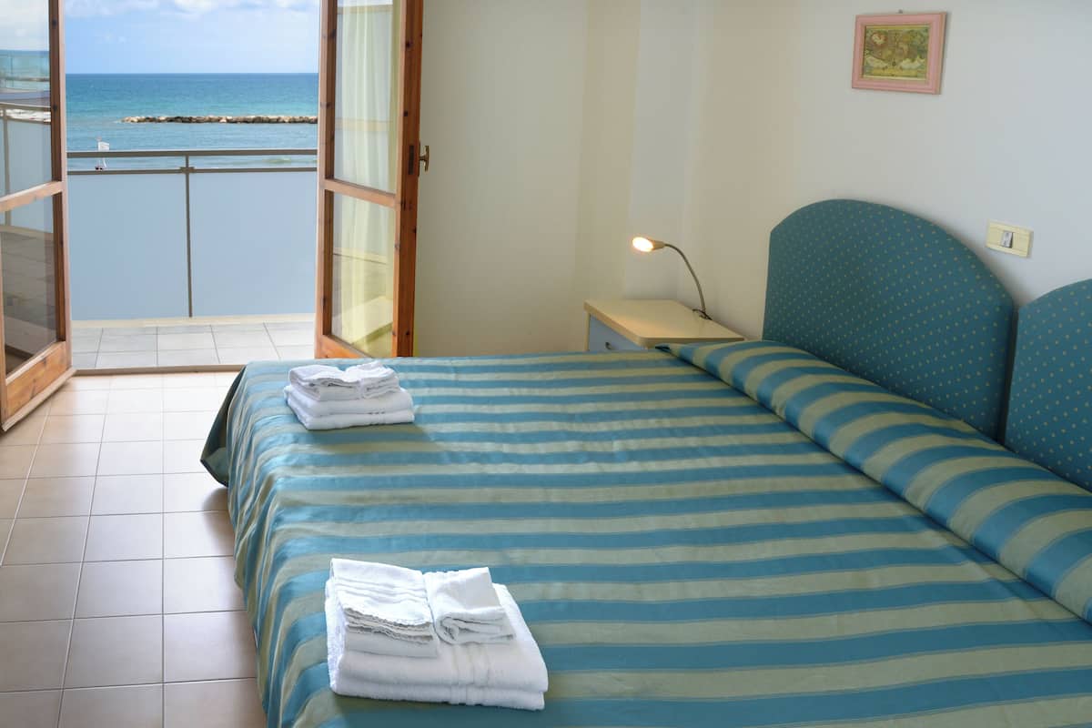 A bedroom with a bed, a nightstand, a lamp, and a view of the sea through an open door.